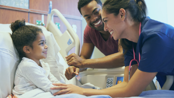 A clincian smiles with a young child sitting in a hospital bed.